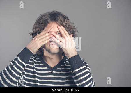 Porträt der jungen hipster Mann in Pullover lugen in die Kamera durch seine Finger. Bild von peinlich männlichen Blick durch seine Hände, die seine Stockfoto