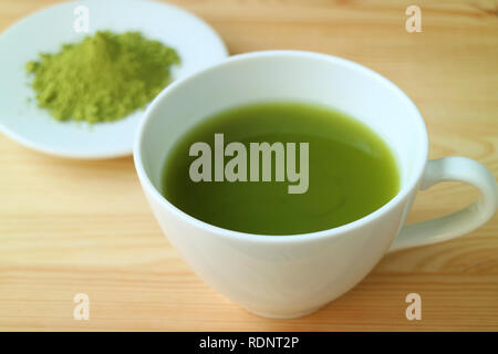Geschlossen, eine Tasse heißen Matcha grüner Tee serviert auf Holztisch mit verschwommenen Platte von Matcha Tee Pulver im Hintergrund Stockfoto
