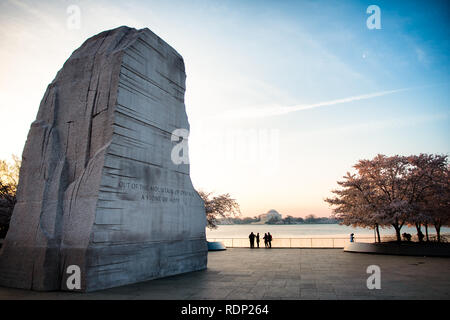 WASHINGTON DC – Kirschblüten blühen bei Sonnenaufgang in der Nähe des Martin Luther King Jr. Gedenkstätte am Tidal Basin. Die Granitskulptur „Stone of Hope“ des Gedenkwerks mit der Inschrift „Out of the Mountain of Desair, a Stone of Hope“ ist mit dem Jefferson Memorial in der Ferne über dem Wasser sichtbar. Stockfoto
