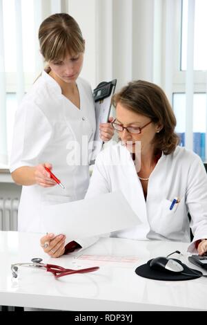 Die medizinische Praxis, Arzt Untersuchung der Gesundheitsdaten eines Patienten an ihrem Schreibtisch, Sie diskutieren mit einem Assistenten Stockfoto