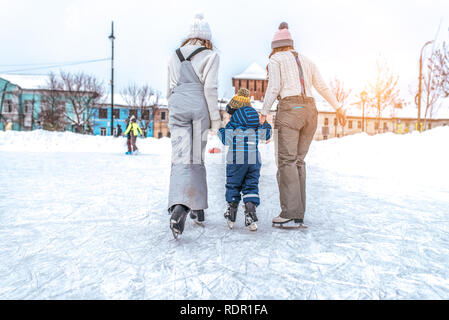 Zwei Freundinnen fahrt mit einem kleinen Jungen 3-5 Jahre alt. Im Winter, in der Stadt, Ausbildern ihren jungen Sohn auf Schlittschuhen zu stehen. Stockfoto