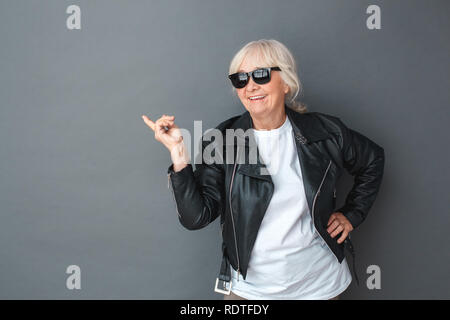 Ältere Frau in Lederjacke und Sonnenbrille Studio stehen isoliert auf Grau zeigt neben freudigen Stockfoto