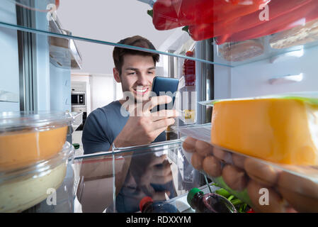 Glücklicher Mann stand vor der Kühlschrank mit Mobilephone In der Küche Stockfoto