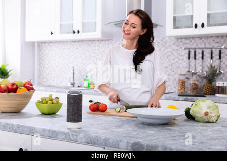 Glückliche Frau im Gespräch mit Voice Assistant beim Schneiden Gurken mit Messer Stockfoto