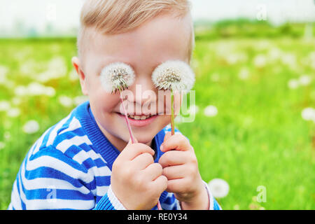 Happy Face kid Lächeln. Close up portrait Freude Kind im Freien. Kleinen Jungen verspielten Lächeln holding Löwenzahn auf Augen, als Brillen. Frohe Kindheit, Sommertag. Hintergrund grüne Gras. Stockfoto