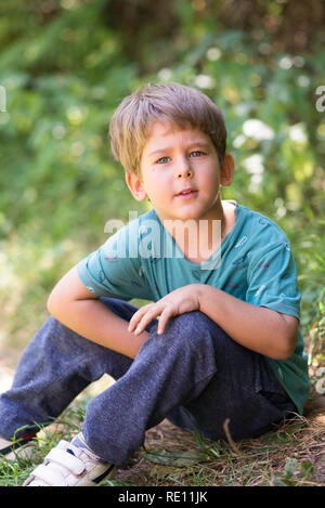 Ein Junge sitzt auf dem Gras, genießt den heißen Sommertag in der Natur, lächelt und spricht neugierig fröhlich Stockfoto