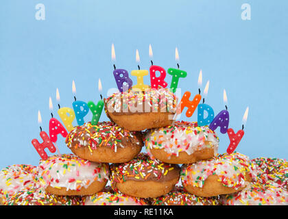 Schokolade und weißem Milchglas Donuts in Candy Streuseln bedeckt in einem Kuchen Stapel mit Happy Birthday Kerzen hell brennen angehäuft. Blauen Hintergrund. Stockfoto