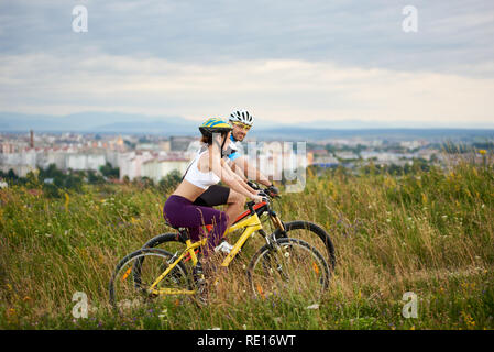 Zwei positive und energetische Radfahrer reiten Fahrrad auf Trail im hohen Gras. Schön und freundlich der Mann und die Frau in der Sportswear und Helme. Hinter ihnen Stadt und der gebauten Umwelt. Stockfoto