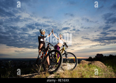 Zwei fröhlich und positiv Paar Radfahrer highing Fünf, lächelnd und an jedem anderen suchen. Schöne Frau und robusten Mann, Sportswear, Helme und Sport Brille, auf Rock Hill posieren. Stockfoto