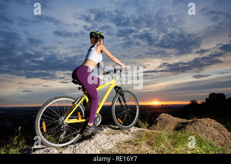 Seitenansicht der athletische und sportliche Mädchen sitzen auf Ihrem Fahrrad und halten die Hände auf. Schlanke weibliche Radfahrer auf Hill posiert mit herrlichem Blick auf die Landschaft und den Sonnenuntergang. Stockfoto