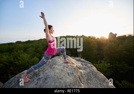 Attraktive schlanke Frau zu tun stretching Yoga Übungen oben auf der großen Felsen auf Green Tree Tops und klaren Himmel im Hintergrund. Sport, Yoga, Fitness und gesunde Lebensweise. Virabhadrasana Stockfoto