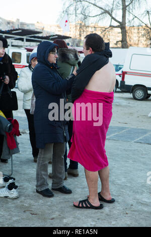 Frau in grauen Jeans und Wintermantel hilft einem Mann in rosa Handtuch, Hausschuhe am Ufer nach Winter orthodoxen Taufe stürzen in Dnepr Fluss im Januar. Stockfoto