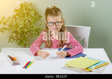 Ein jüngeres Schulmädchen mit Brille schreibt etwas mit der linken Hand auf dem Notebook- und setzt sich an den Tisch. Zurück zum Konzept der Schule. Das Kind ist links Stockfoto