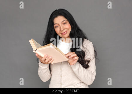 Freestyle. Reife Lady stehen isoliert auf Grau lesen Buch neugierig Stockfoto