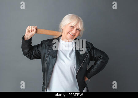 Ältere Frau in Leder Jacke Studio stehen auf Grau mit Rolling Pin auf der Schulter isoliert entspannt Stockfoto