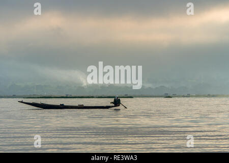 Ein einsamer Fischer an der Spitze seines Bootes gehockt, als er am Rande der Inle See, Myanmar mit einem Smokey und bewölkt Hügel im Hintergrund Fische. Stockfoto