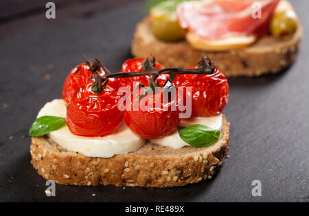 Nahaufnahme von Bruschetta mit Frischkäse, Cherry Tomaten, Basilikum, geräucherter Käse, Schinken, Oliven auf einen hölzernen Tisch. Stockfoto