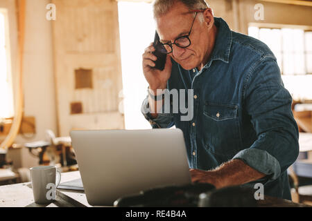 Reifen Mann, besetzt mit Laptop und Gespräch am Handy in seiner Werkstatt. Ältere männliche Tischler arbeiten, die auf Notebook- und Anruf in seinem carpen Stockfoto