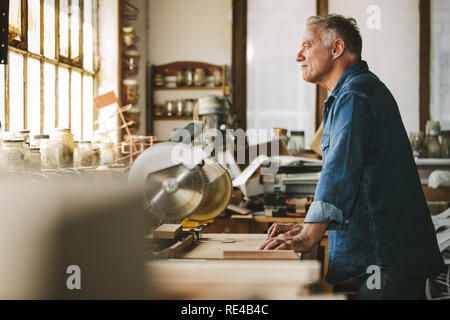Seitenansicht des Senior Zimmermann steht an der Werkbank und Wegsehen. Nachdenklich männliche Tischler in seiner Werkstatt. Stockfoto