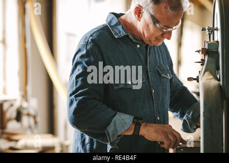 Reife männliche Zimmerei Arbeitnehmer die Arbeit in seiner Werkstatt. Tischler Schneiden von Holz auf Band Maschine sah in der Werkstatt. Stockfoto