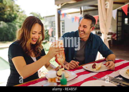 Lächelnd Mann und Frau essen Hot Dogs sitzen am Esstisch. Frau gießen Tomatensauce in ihren Teller beim Essen. Stockfoto