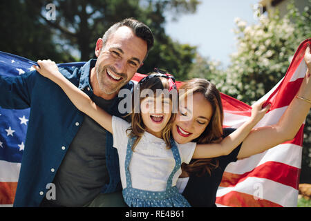 Lächelnd Mann und Frau mit einem Kind stehen im Freien halten Sie die amerikanische Flagge hinter Ihnen. Glückliche Familie zusammen im Freien halten der amerikanischen Stockfoto