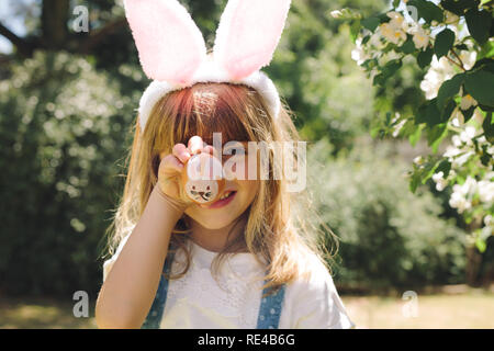 Nahaufnahme von einem lächelnden Mädchen, dass eine bemalte Ostereier vor ihrem Gesicht. Mädchen mit Hase ohr-kopfband Spaß spielen mit gemalten Esser e Stockfoto