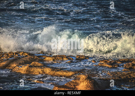 Wellen mit Spray auf Felsen am Ufer bei Sonnenuntergang Stockfoto
