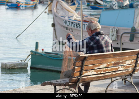 Marsaxlokk/Malta - 30. November 2018: Ein alter Fischer ist die Reparatur seiner Fischernetze auf einer Bank am Hafen. Stockfoto