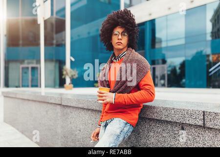 Junge curly Mixed-race Junge trinkt Kaffee auf der Straße vor dem Hintergrund von Bürogebäuden. Jugend Kultur und Mode. Student auf eine Pause zwisch Stockfoto