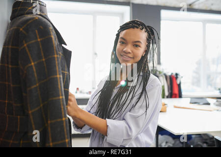 Junge weibliche Maßgeschneiderte stehen in der Nähe der Puppe mit Jacke gekleidet und lächelnd im Atelier Stockfoto