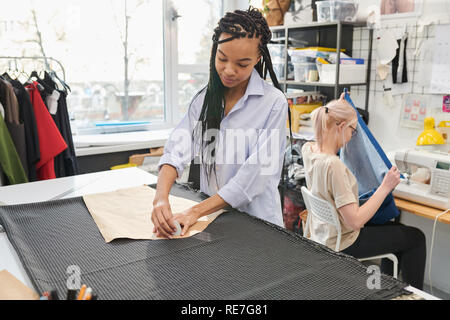 Junge Schneider arbeiten mit Stoff und Muster, während ihr Kollege mit Nähmaschine auf dem Hintergrund in der Werkstatt Stockfoto