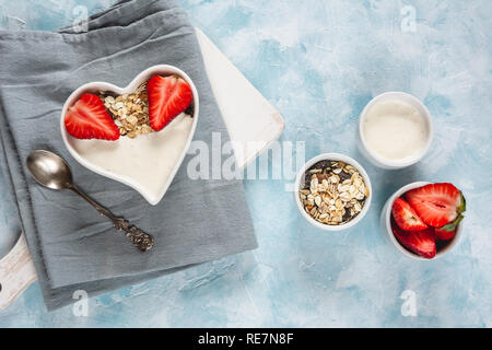 Joghurt mit Müsli und Erdbeeren in eine Herzförmige Schale auf einem Pastell-blaue Hintergrund. Stockfoto