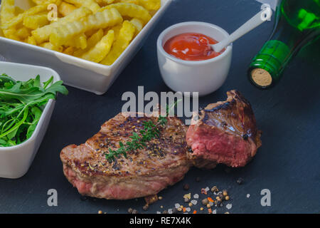 Gegrilltes Steak mit Pommes Frites und auf schwarzen Steinplatte auf hölzernen Tisch serviert. Stockfoto