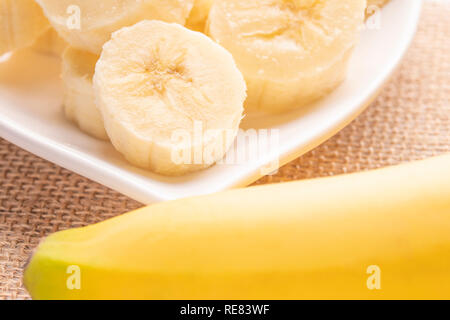 In Scheiben geschnitten und ganze Bananen auf einem weißen herzförmigen Platte und einem alten Holztisch in den Strahlen der Sonne. Wicker Serviette. Diät gesetzt Stockfoto