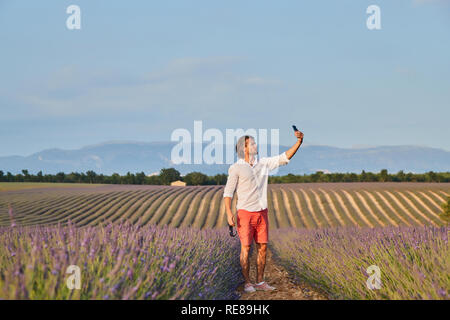 Die schöne brutale jungen Mann mit langen brünetten Haar macht selfie per Handy im Bereich der Lavendel, weiße Hemden und rote Shorts Stockfoto