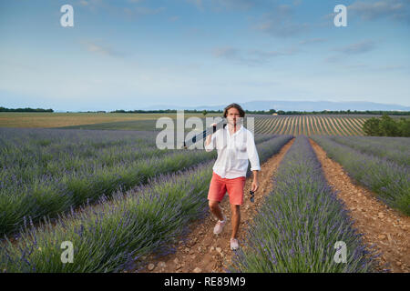 Die schöne junge brutalen Mann geht zu einem Lavendelfeld bei Sonnenuntergang, er ist in ein weißes Hemd mit kurzen Ärmeln und rote Shorts gekleidet, der Fotograf Stockfoto