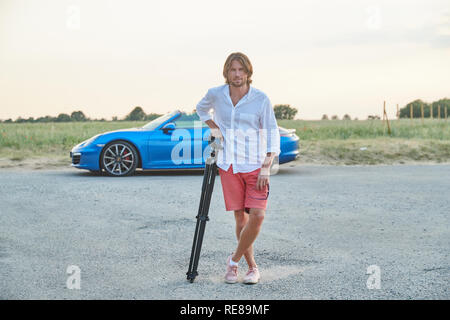 Die schöne junge brutalen Mann steht auf einem blauen Sportwagen bei Sonnenuntergang, er ist in ein weißes Hemd mit kurzen Ärmeln und rote Shorts gekleidet Stockfoto