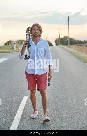 Die schöne junge brutalen Mann steht auf der Straße bei Sonnenuntergang, er ist in ein weißes Hemd mit kurzen Ärmeln und rote Shorts gekleidet, der Fotograf Stockfoto