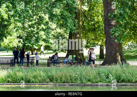 London, UK, 21. Juni 2018: St James Park, grüne Bäume im sonnigen Sommer mit vielen Menschen zu Fuß auf dem Bürgersteig durch Teich Wasser des Flusses und sitzen auf Bänken Stockfoto