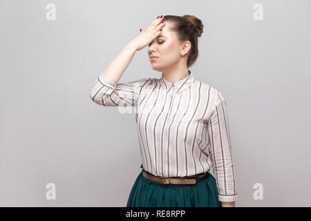 Portrait von unglücklichen traurig schöne junge Frau in gestreiften Hemd und grün-Rock mit gesammelten Verbot Frisur, stehen und die Hand auf die Stirn. Stockfoto