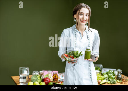 Porträt einer jungen Frau Ernährungsberaterin in Medical Gown stehend mit Salat und Smoothie auf dem grünen Hintergrund im Innenbereich Stockfoto