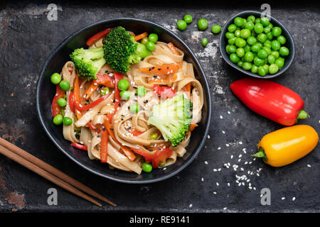 Asiatische Pfannengerichte mit udon Nudeln und Gemüse. Brokkoli, Erbsen, Paprika, Karotten und Pasta in schwarz Schüssel. Ansicht von oben Stockfoto