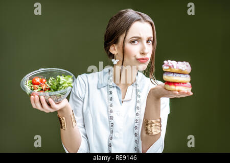 Junge Frau Ernährungsberaterin auf die Krapfen mit traurigen Emotionen Wahl zwischen Salat und ungesunden Nachtisch auf dem grünen Hintergrund Stockfoto