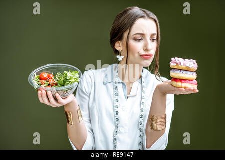 Junge Frau Ernährungsberaterin auf die Krapfen mit traurigen Emotionen Wahl zwischen Salat und ungesunden Nachtisch auf dem grünen Hintergrund Stockfoto