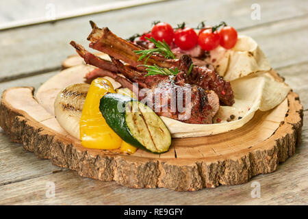 Lamm schrumpfte mit Beilage auf Holzplatte. Lamm Bein mit Garnierung auf Holzplatte. Stockfoto