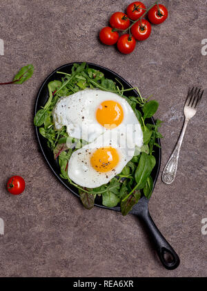 Zwei Spiegeleier auf Pan mit verschiedenen Arten von Kräutern, Rucola, Spinat, Mangold und Cherry Tomaten, mit Gabel auf braunem Hintergrund, Konzept einer gesunden Stockfoto