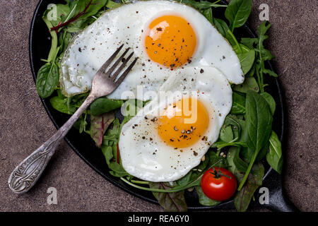 Zwei Spiegeleier auf Pan mit Rucola, Spinat und Tomaten, mit Gabel an der braunen Hintergrund, Konzept der gesunden Frühstück, Nahaufnahme, Makro. Top vie Stockfoto