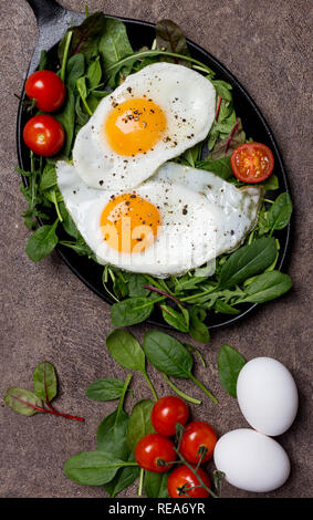 Spiegeleier mit frischen Kräutern und Kirschtomaten im Pan zum Frühstück, auf braunem Hintergrund. Konzept der leckeren und gesunden Lebensstil Stockfoto