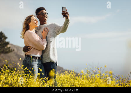 Liebevolle junge Paare auf Sommer Urlaub nimmt ein Selbstbildnis mit Smartphone im Freien. Entspannt junges Paar auf Feiertag selfie stehend in Mead Stockfoto